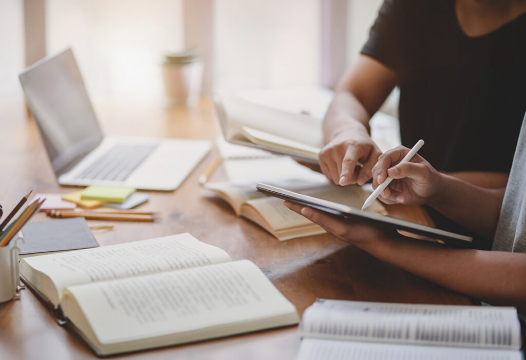 Two people collaborating on a tablet beside books and a laptop.